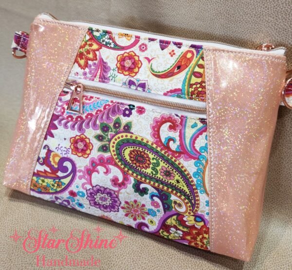 Stunning "Just Peachy" Sunshine Crossbody Crossbody. Measures 10 1/2" wise x 8" tall and 3" deep. $85.00 Colorful paisley floral fabric center, with striking holographic peach glitter vinyl. Rose gold hardware really ins striking in this combination. The exterior back also has a sipper pocket with rose gold zipper teeth and zipper pull. Matching adjustable strap in paisley and can be worn as a side strap or crossbody. Interior is waterproof canvas in rose gold color. It as a roomy slip pocket and zipper pocket.